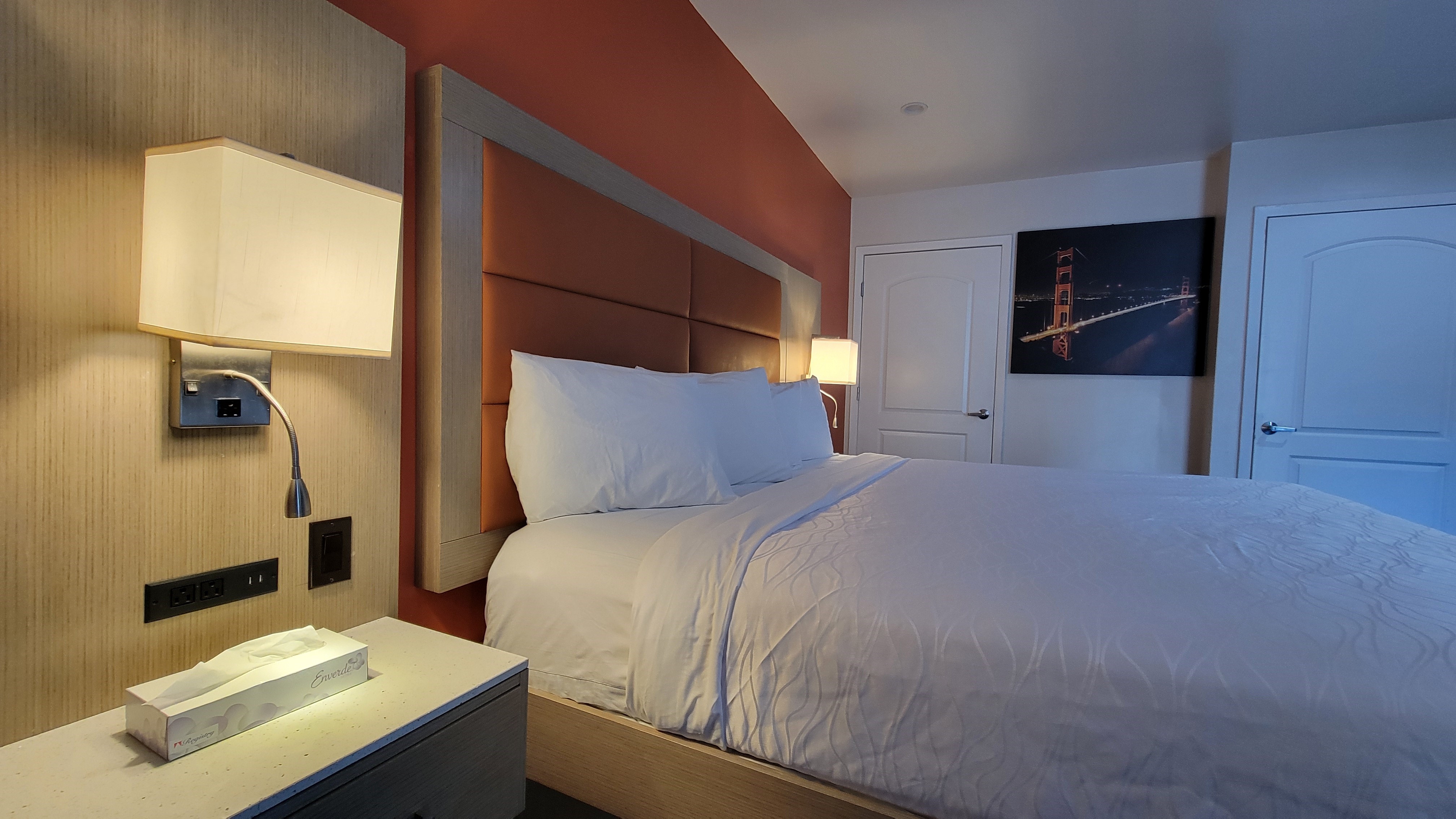 A hotel room with a bed, nightstand, lamp, and artwork on the wall.