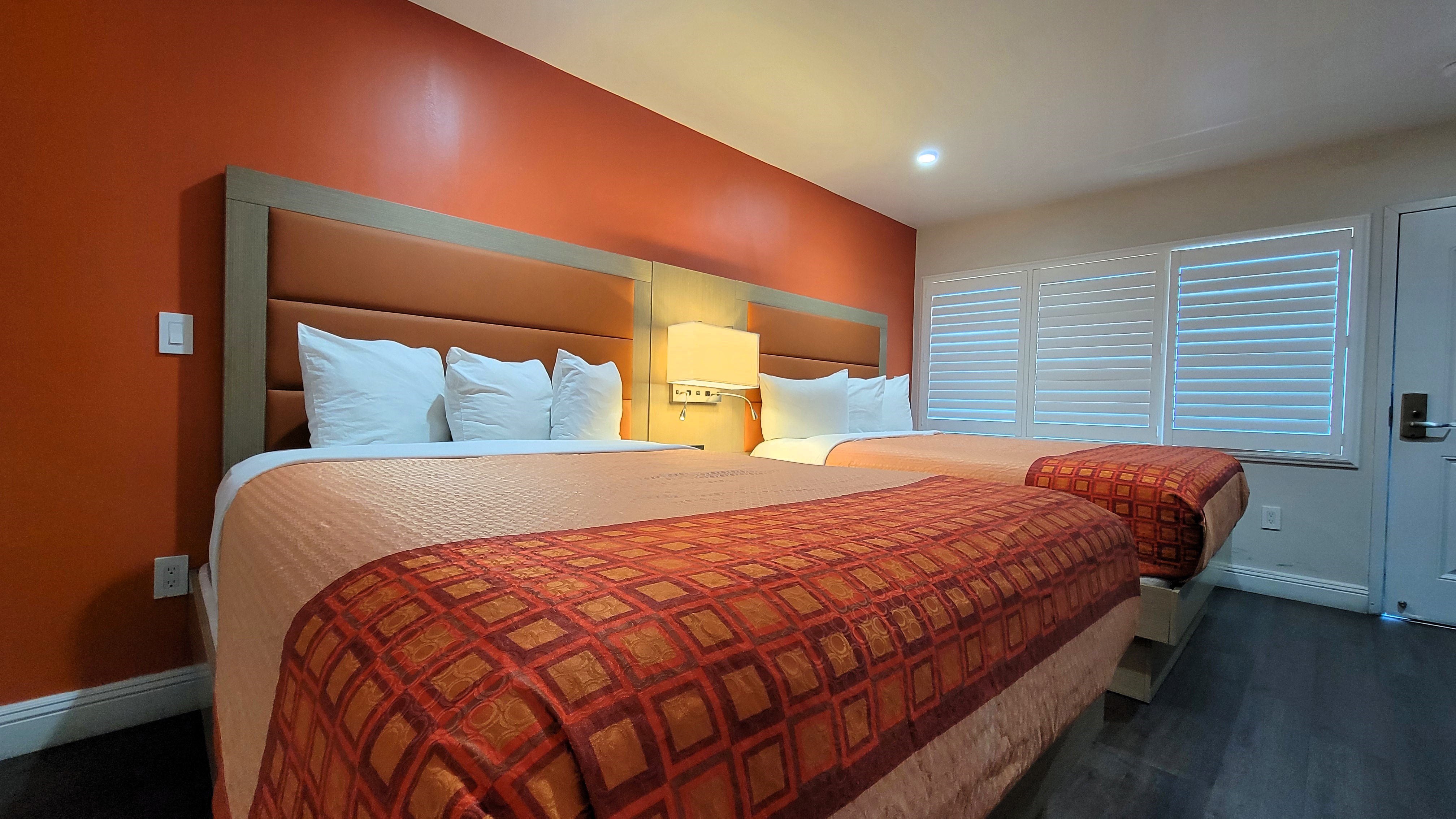 A hotel room with two beds, an orange accent wall, and a window with blinds.