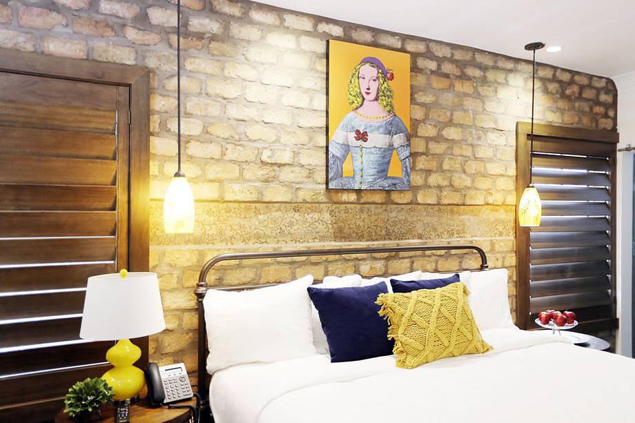 A bed with white linens and decorative pillows is positioned under a painting on a brick wall, with pendant lights and a telephone nearby.