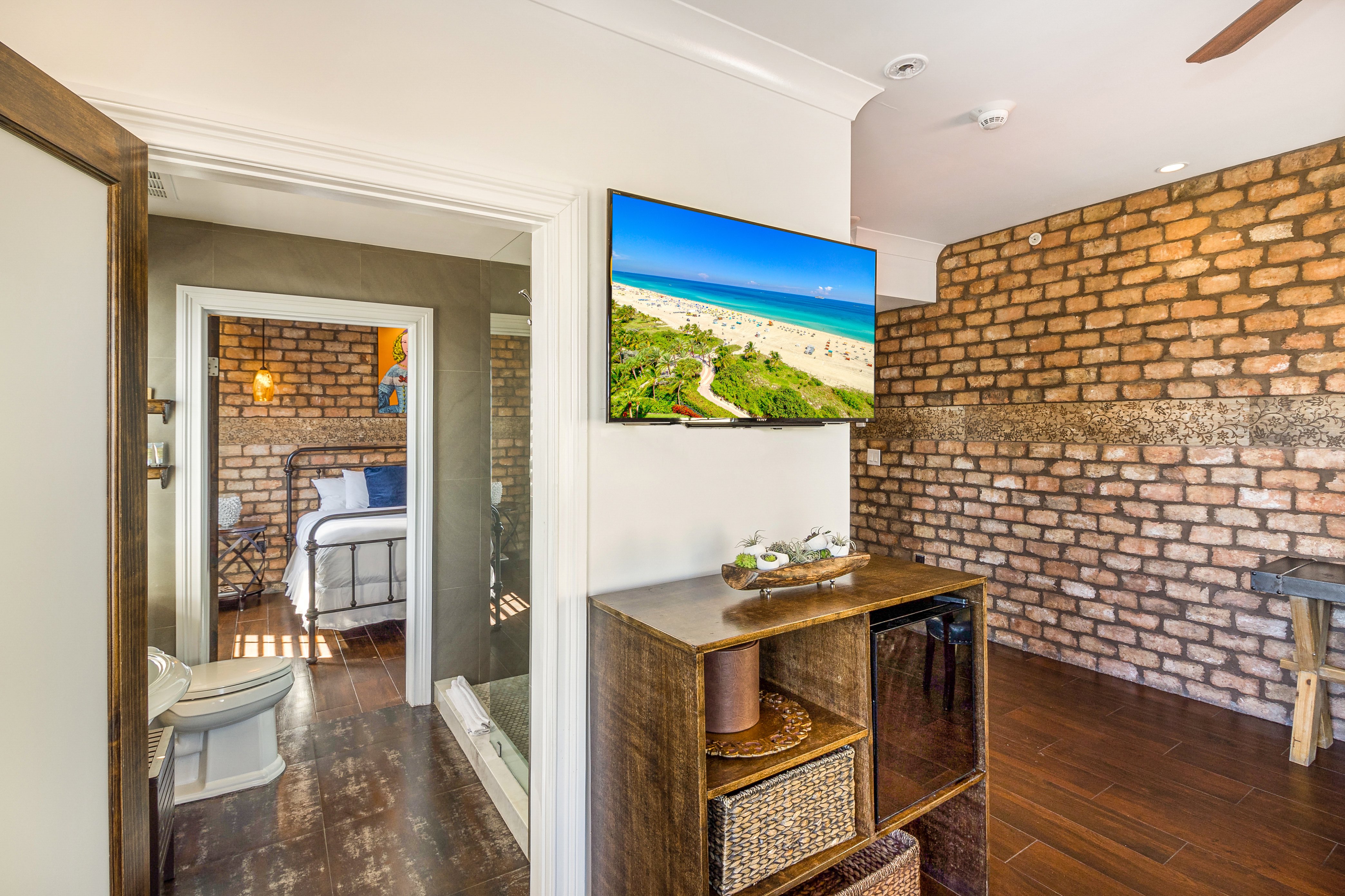 A hotel room with a TV on the wall, a wooden shelf, and a doorway leading to a bedroom with a bed.